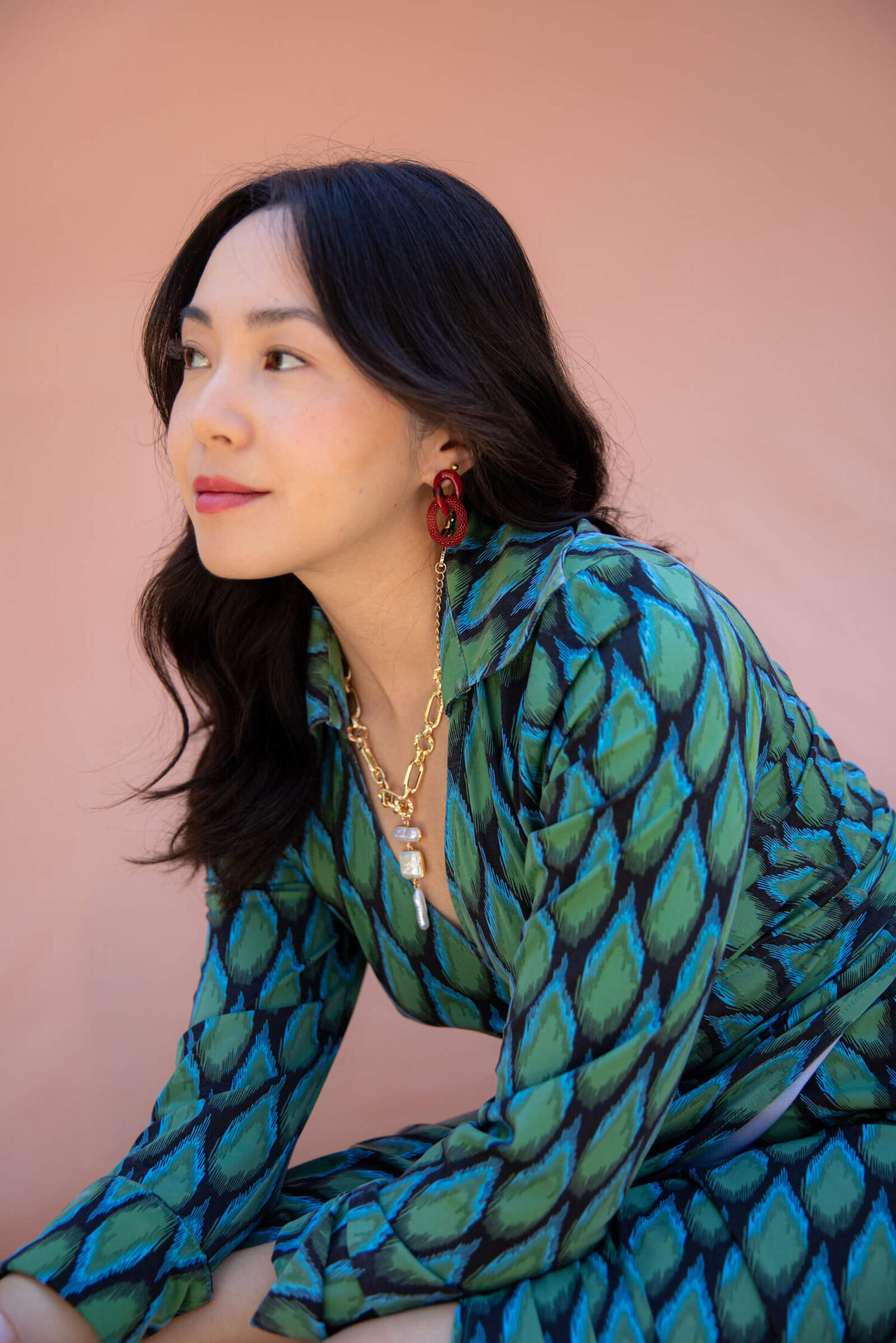 portrait of the author looking into the distance. she is sitting and wearing a green wrap dress, gold chain, and red earrings