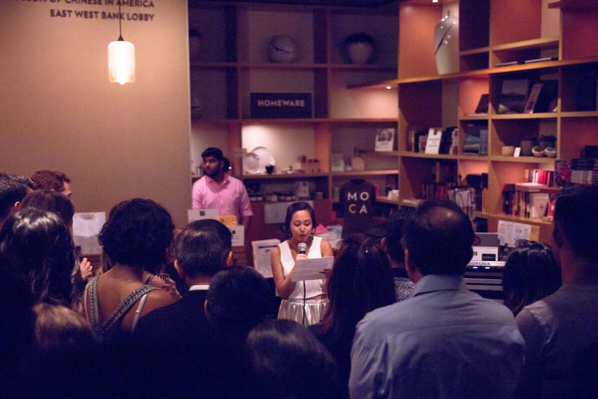 Photo of the author reading into a microphone as an audience looks on